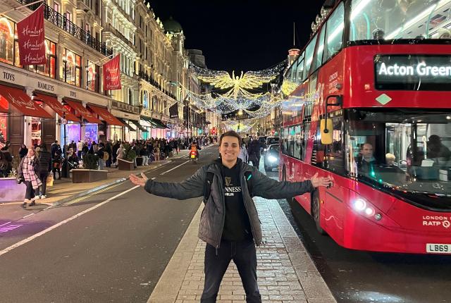 london student standing in street with arms stretched out next to red bus and string lighted buildings