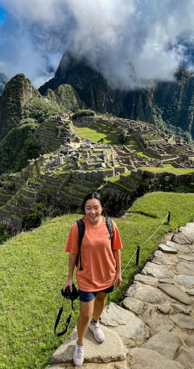 a student posing for a photo with great views of Machu Picchu and mountains in the background on a blue and cloudy day